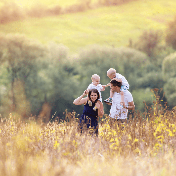 Family with two young boys on their father's shoulders walking through a sunlit meadow