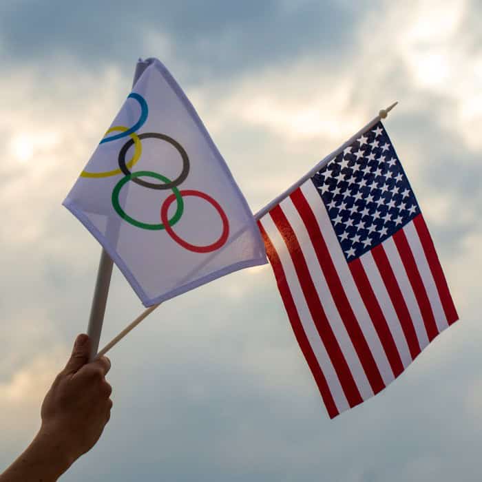 A hand holding an Olympic-rings flag and an American flag against a cloudy sky.