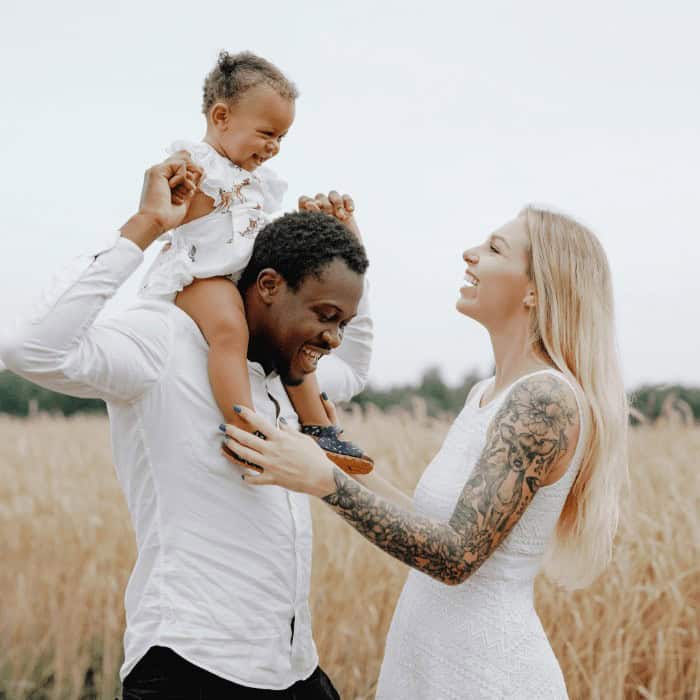 A young multiracial family smiling together in a field, the father lifting a toddler on his shoulders.