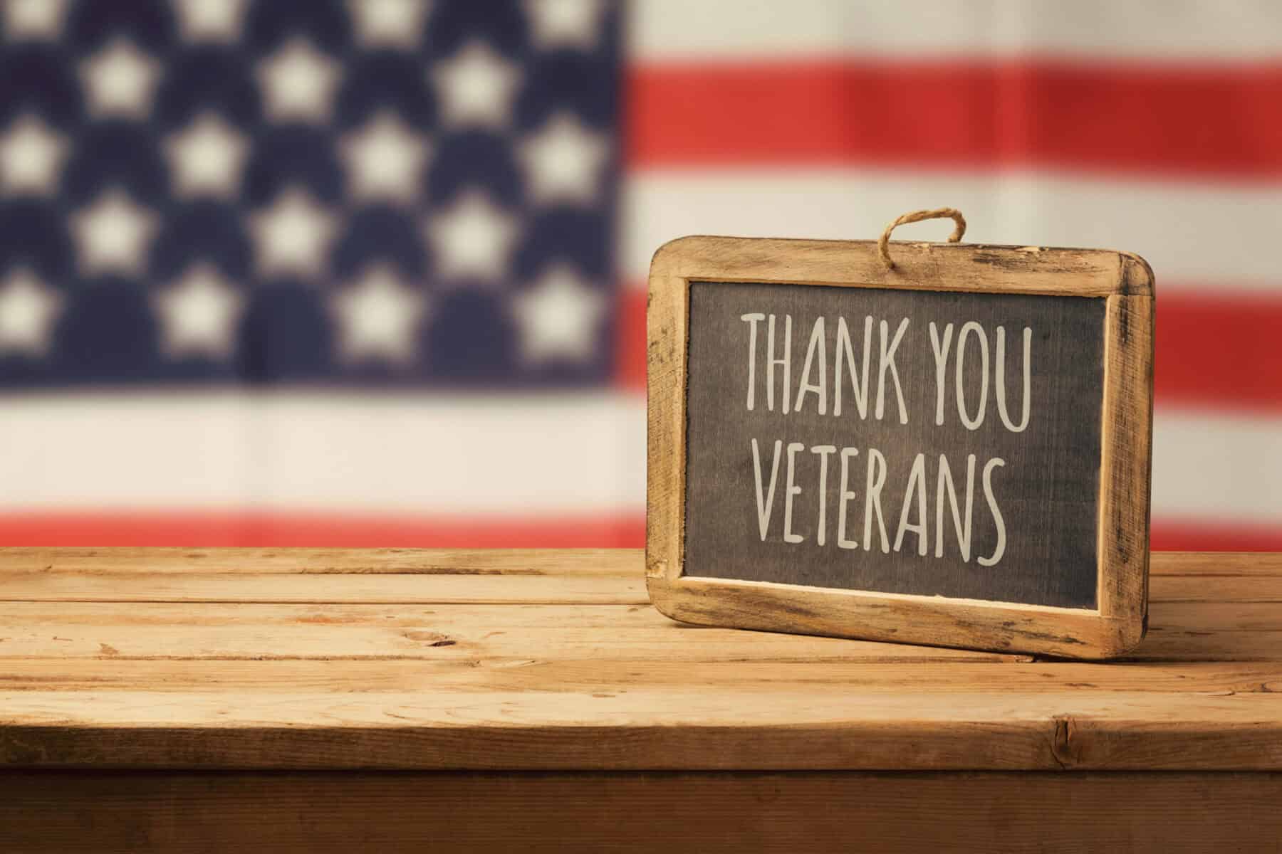 A "Thank You Veterans" chalkboard sign resting on a wooden table in front of an American flag.