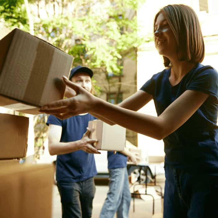 A young woman handing a moving box to two movers in matching shirts outside a building.