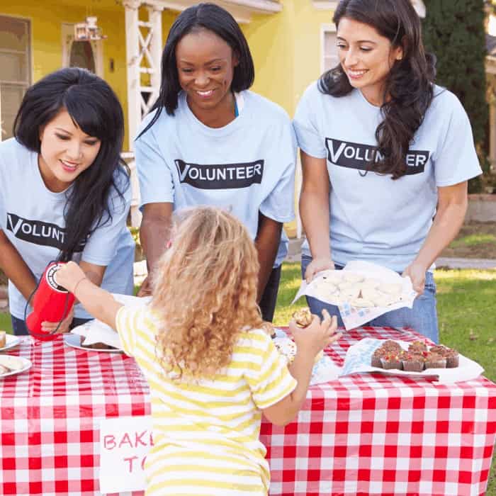 Three volunteers in matching t-shirts serving baked goods to a child at an outdoor charity bake sale.
