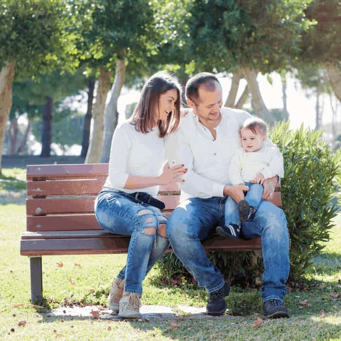 Parents and a young child sitting together on a park bench on a sunny day.