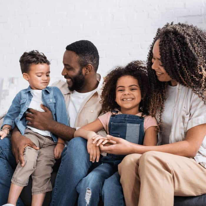 A smiling family of four — two parents with a young son and daughter — sitting together at home.