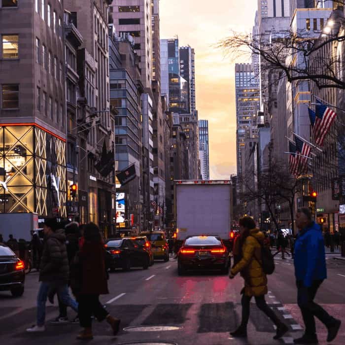 A busy New York City street at dusk with pedestrians, traffic, and American flags on the buildings.