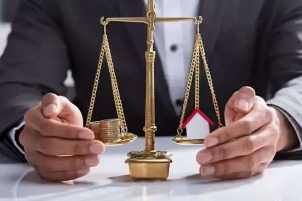 A man in a suit rests his hands either side of a brass balance scale weighing a stack of coins against a small model house.