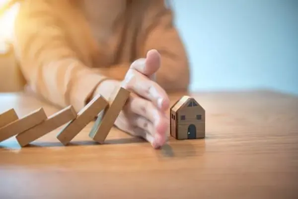 A woman's hand stops a line of toppling wooden blocks before they reach a small wooden house.