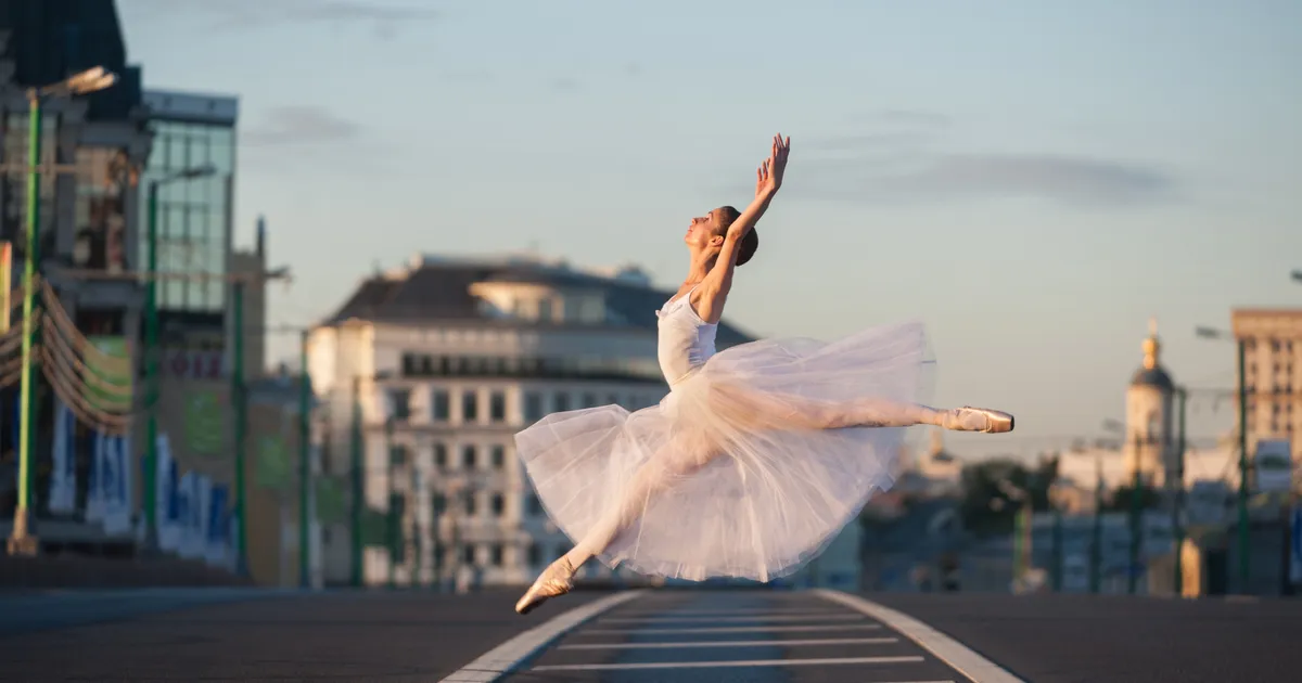 A ballerina in a white tutu leaps gracefully on an empty city street at sunrise.
