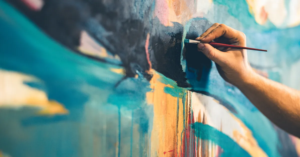 A hand paints an abstract canvas in blue, teal, and orange hues.