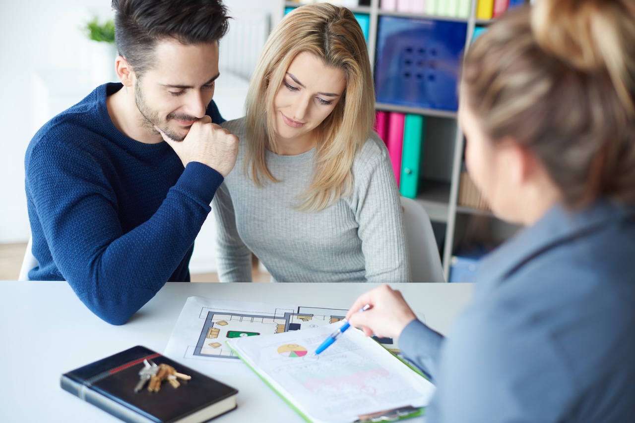 A couple reviews floor plans and financial documents with an advisor across a desk, house keys resting on a notebook nearby.