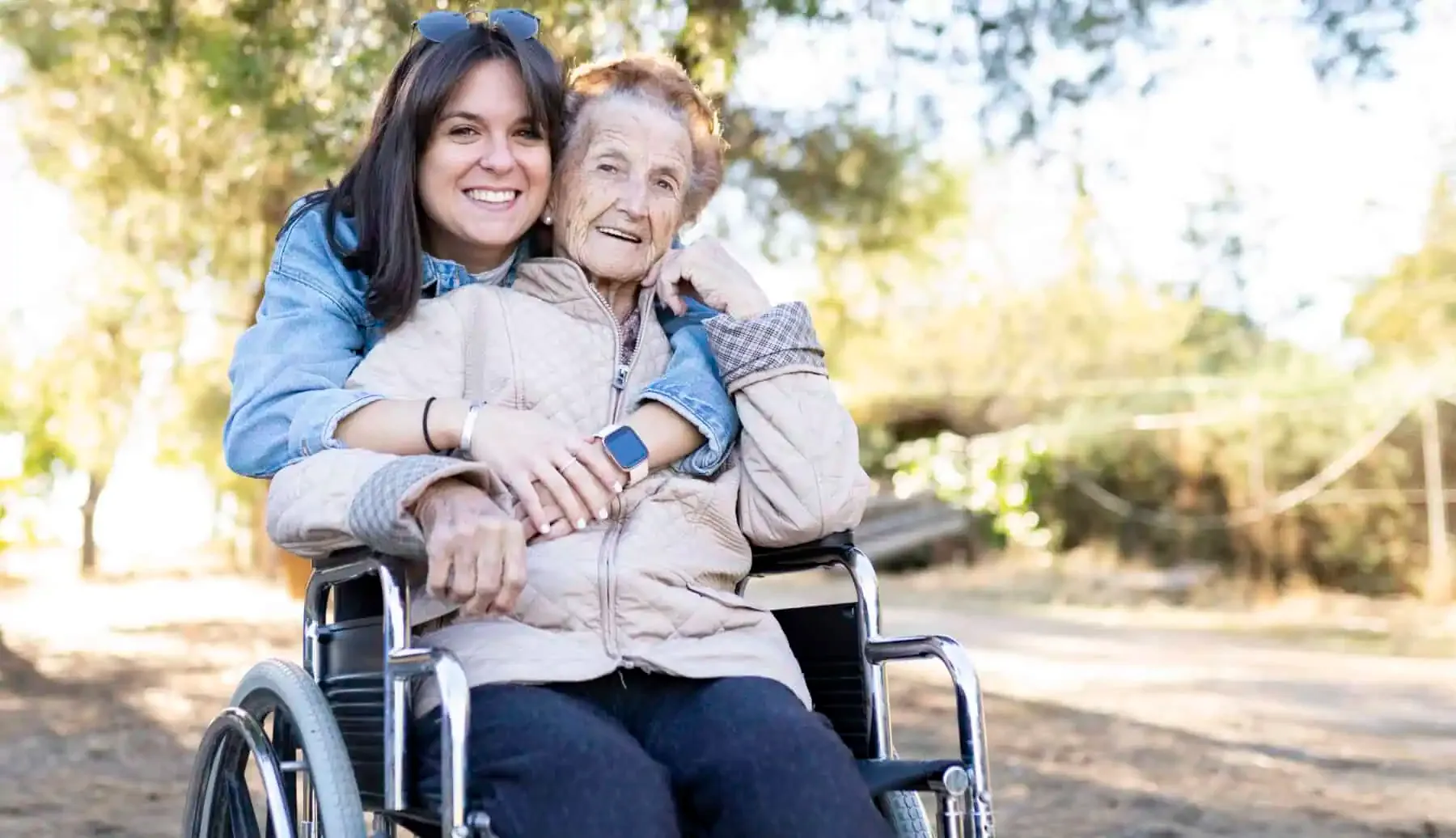 A young woman leans down to embrace a smiling elderly woman seated in a wheelchair on a sunlit tree-lined path.