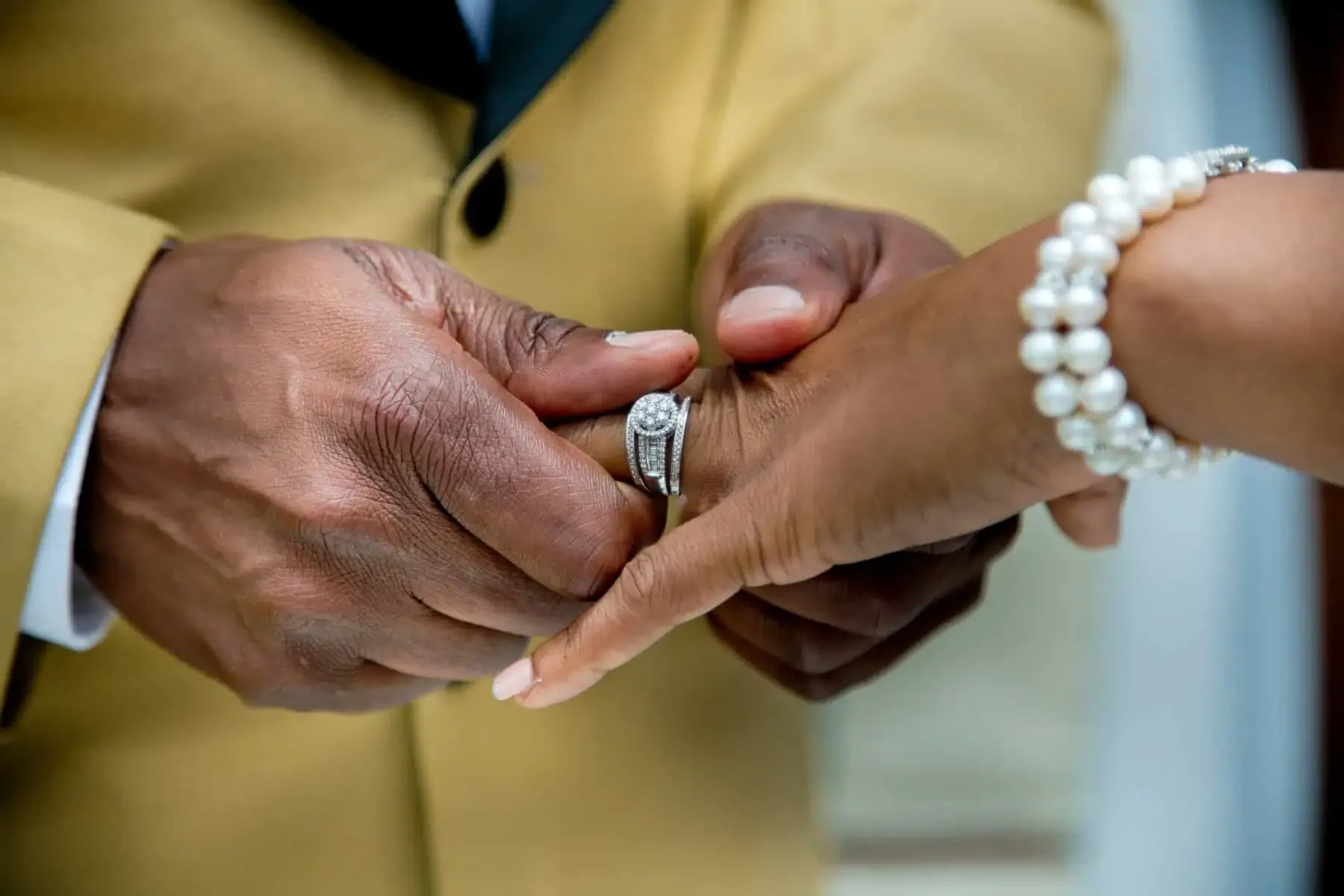 Close-up of a man sliding a diamond ring onto a woman's finger; she wears a pearl bracelet and he a yellow jacket.