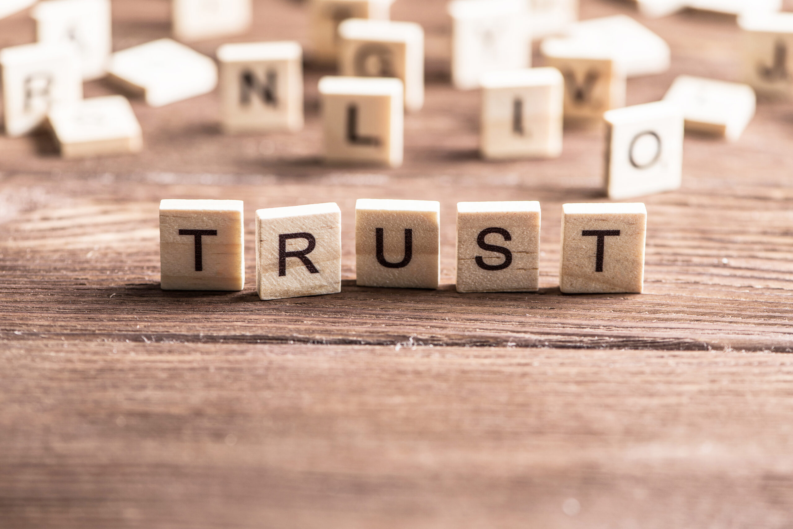 Wooden letter tiles spelling "TRUST" on a weathered wooden table, with scattered tiles blurred behind them.