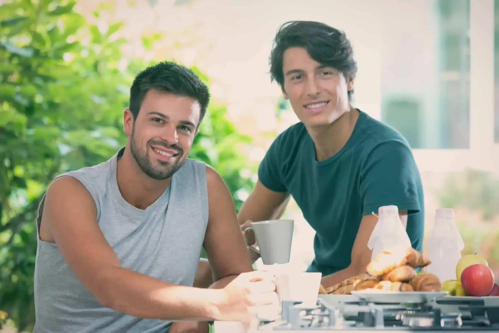 Two smiling men sit together over coffee and pastries at a sunlit breakfast table.
