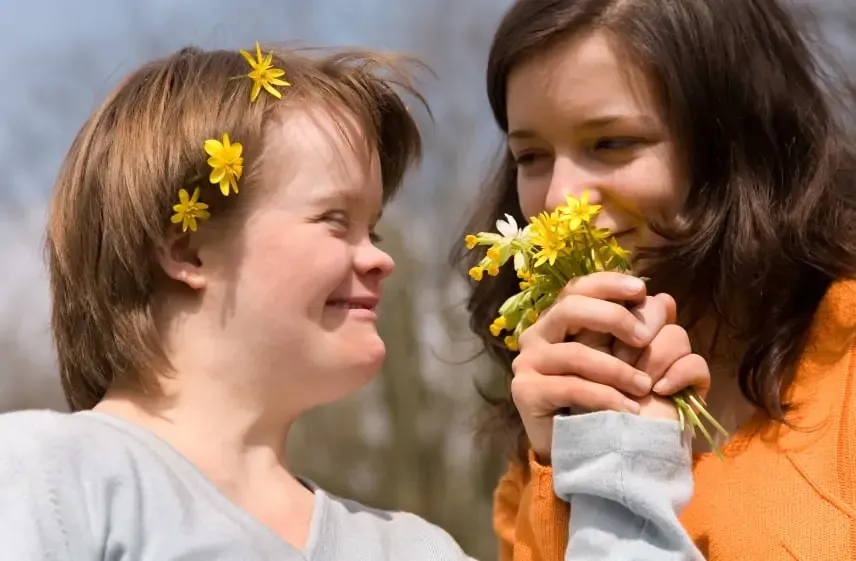 A smiling girl with Down syndrome looks up at another girl who is holding a bunch of yellow spring flowers.