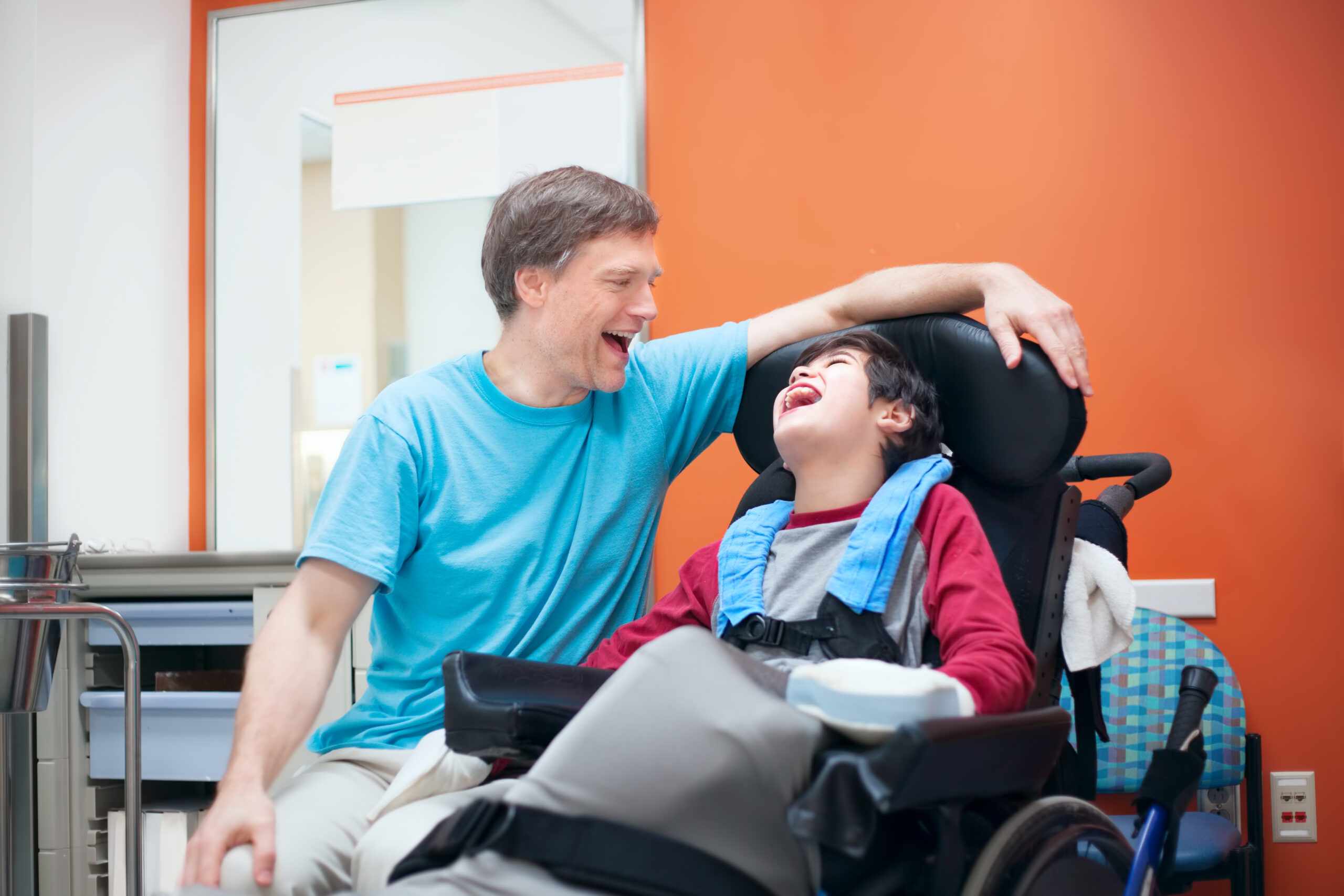 A man laughs with a boy seated in a supportive wheelchair in a clinic room with an orange wall.