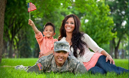 A soldier lying on the grass with his wife and young son, who waves a small American flag, in a sunlit park.