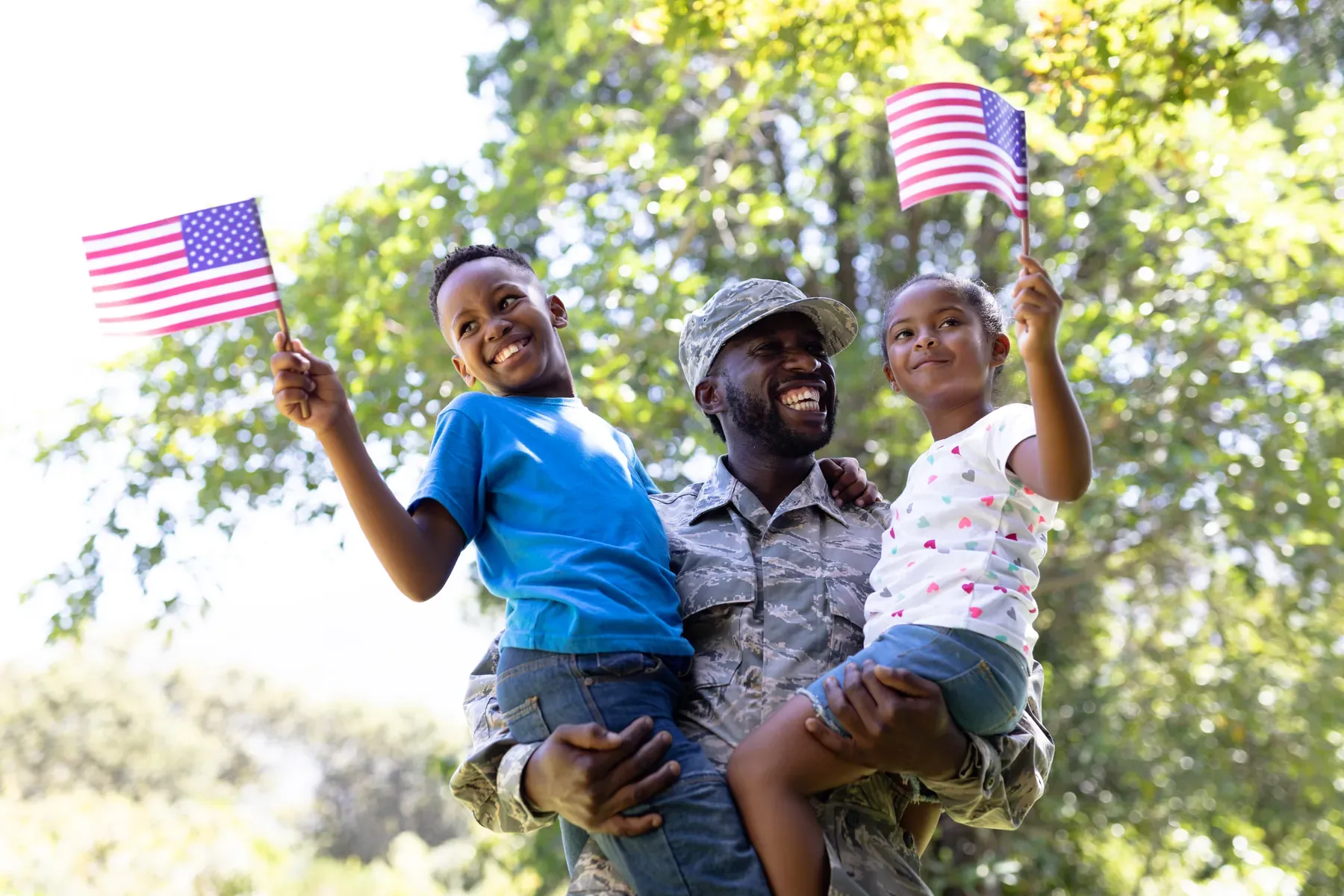 A smiling soldier in uniform holds a young boy and girl, both waving small American flags, beneath sunlit trees.