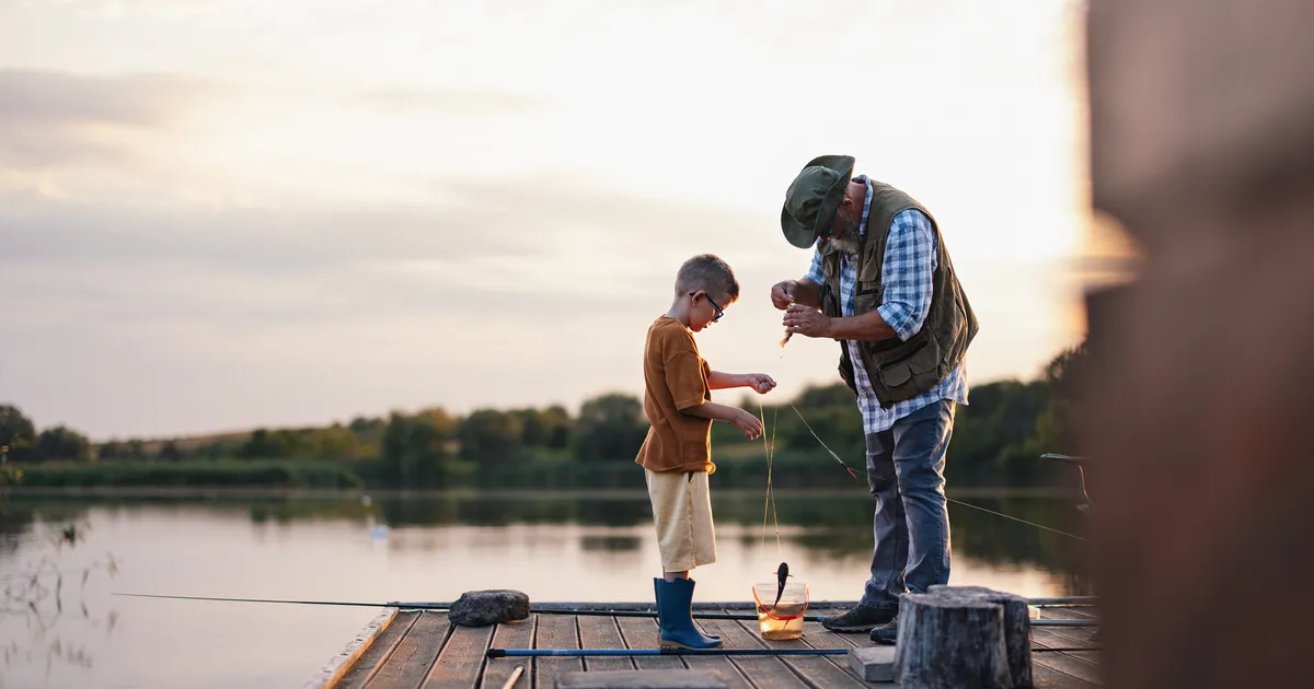An older man and a young boy bait a fishing line together on a lake dock at sunset.