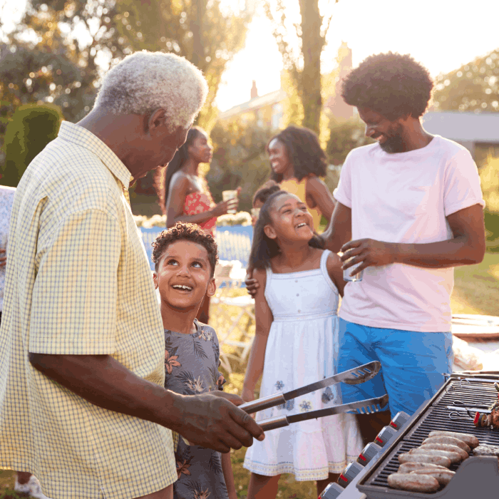 Family enjoying a barbeque with the grandfather at the grill talking to his grandson.